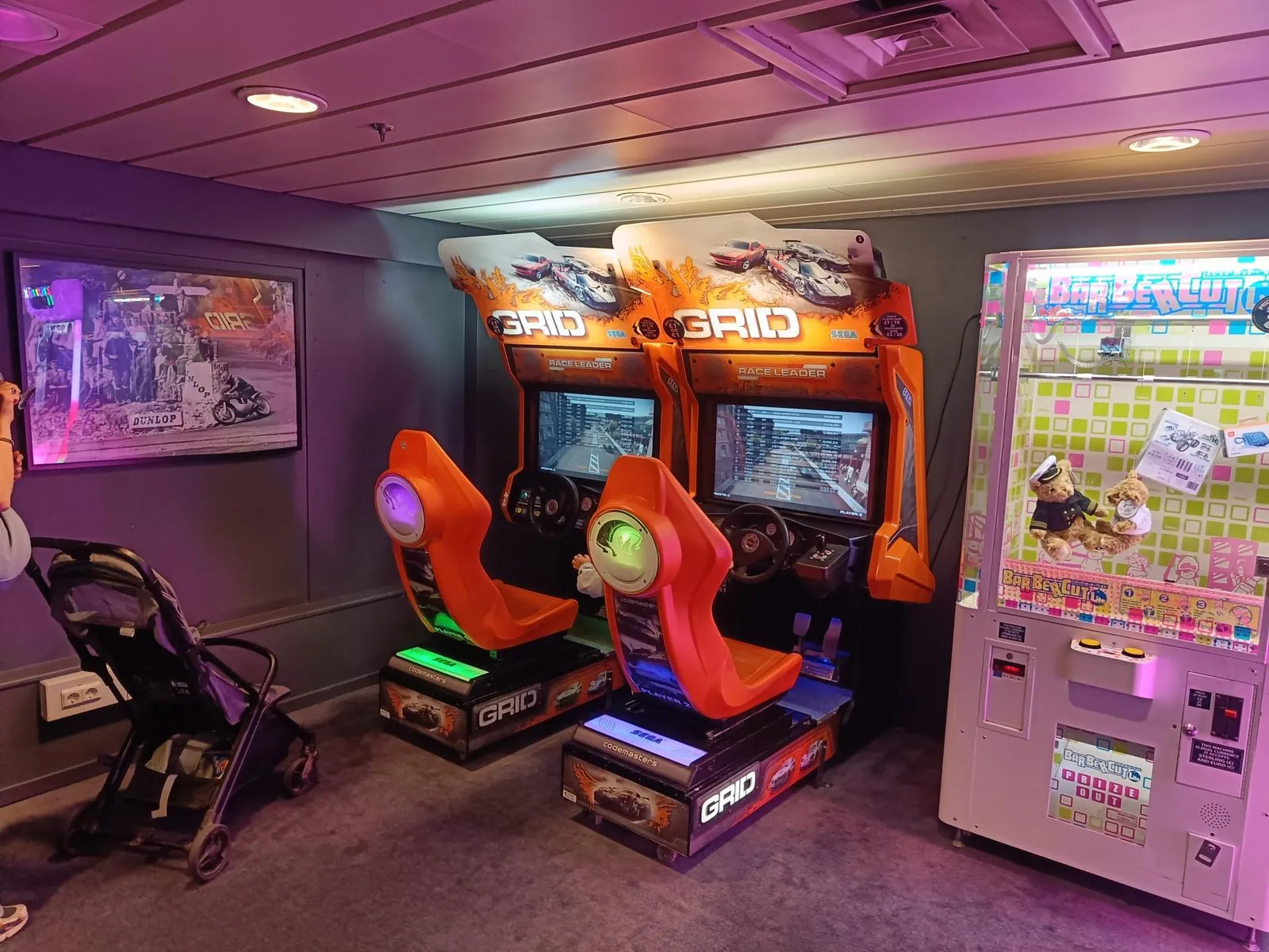 Twee oranje racegamekasten en een grijpmachine in de speelhal aan boord, met een kinderwagen ernaast
