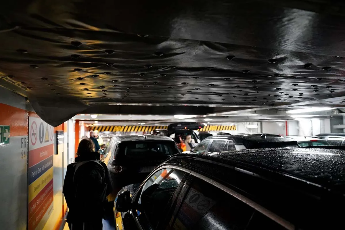 Auto's dicht op elkaar op een autodek met een laag hangend tussendek erboven; passagiers lopen met tassen tussen de auto's door