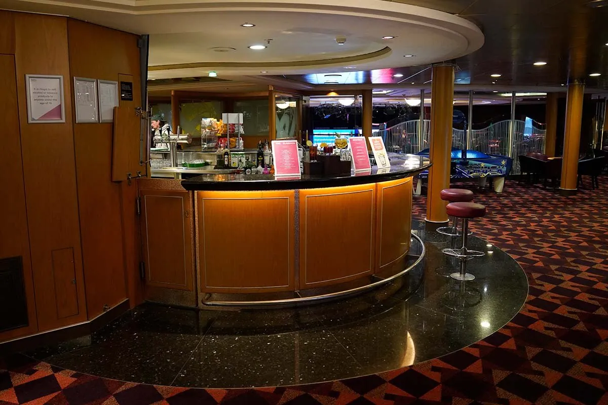 Een ronde houten bar met twee rode krukken op een gepolijste donkere vloer, flessen en menukaarten op de toog en een medewerker erachter; daarachter staan speelautomaten achter een hek
