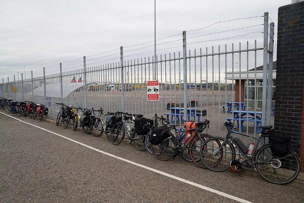 Een rij volgepakte fietsen met bagagetassen tegen een stalen hek bij de terminal van Harwich, met picknicktafels erachter