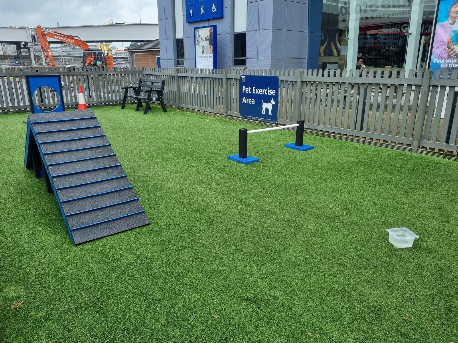 Een uitlaatveld met kunstgras binnen een grijs houten hek, met een hellingbaan, een springlat, een bord met een rond gat, een bankje, een bak water en het bord Pet Exercise Area, voor een terminalgebouw