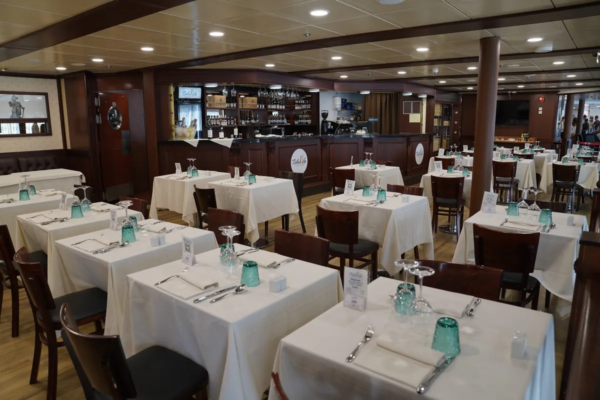Restaurant Dolce Vita aan boord met gedekte tafels met witte kleden en groene glazen, en een houten bar met wijnflessen erachter