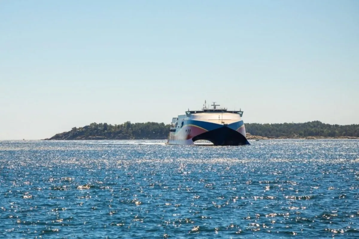 Een snelle catamaranveerboot met een blauw-rode boeg vaart over glinsterend water langs een beboste kust