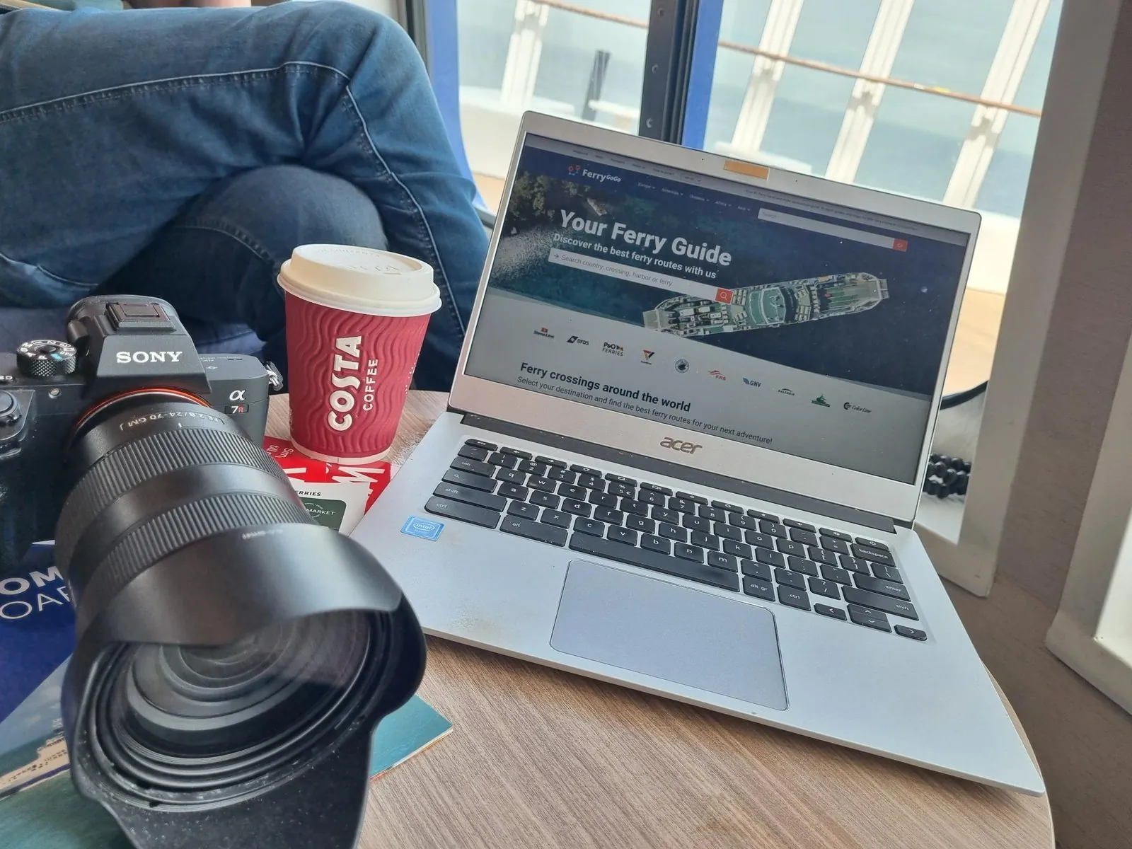 Laptop met de website van Ferrygogo op een tafel bij het raam, met daarnaast een beker Costa-koffie en een fotocamera met telelens