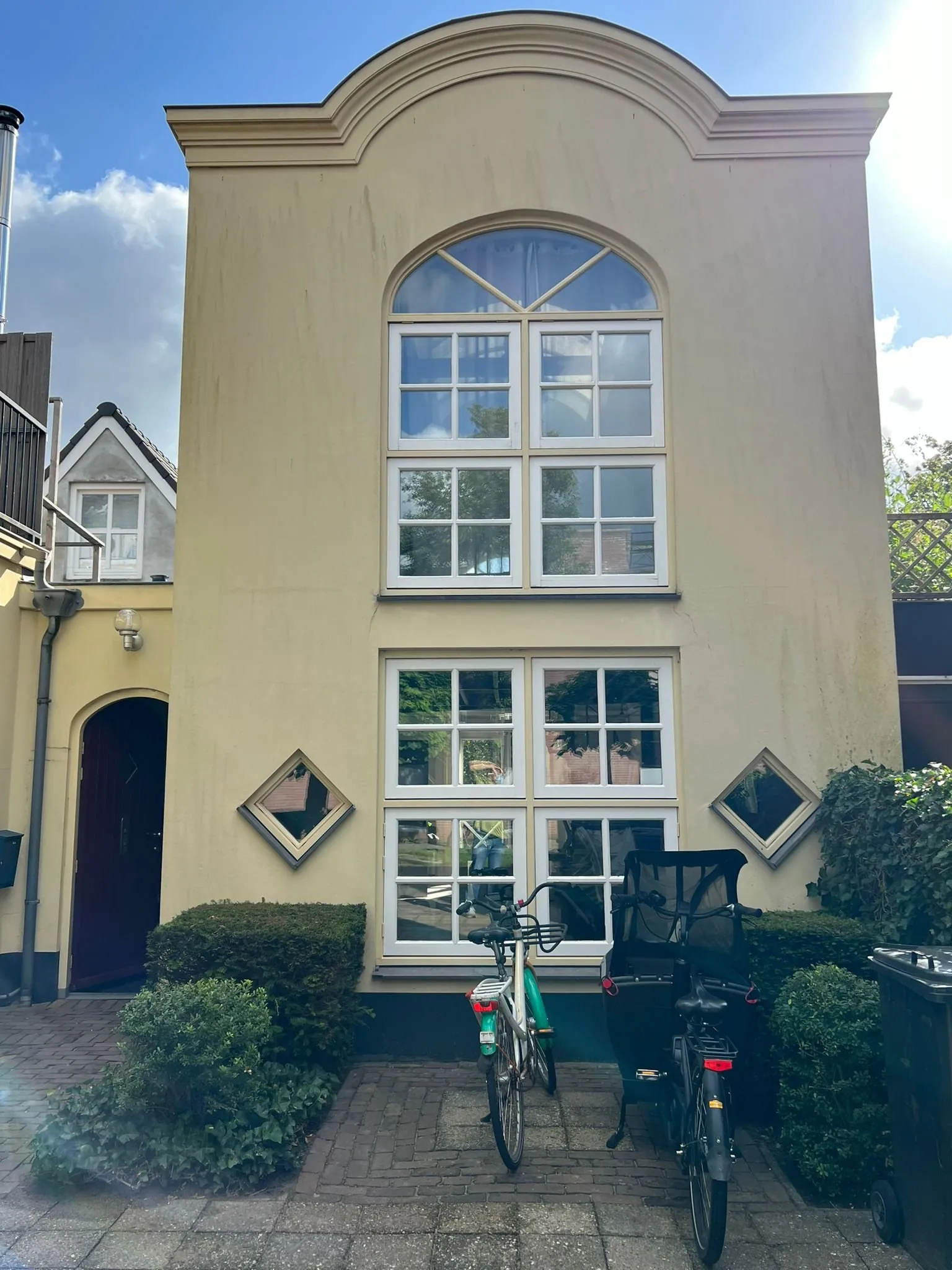 Een geel gepleisterd pand met een boogvormige geveltop, hoge witte roedevensters en een donkerpaarse voordeur, met buxus en twee fietsen op de bestrating ervoor