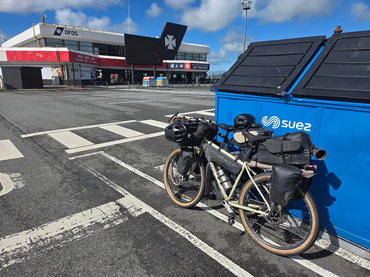 Een fiets met fietstassen tegen een blauwe container op het terrein van de DFDS-terminal, met het terminalgebouw op de achtergrond