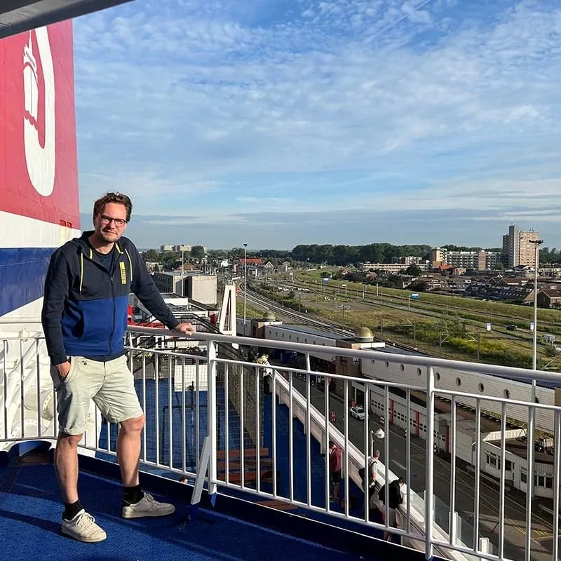 Jelle van der Bij met bril, blauwe hoodie en korte broek op het buitendek van een veerboot, leunend op de reling, met de rode schoorsteen links en een havenstad met spoorlijnen onder hem
