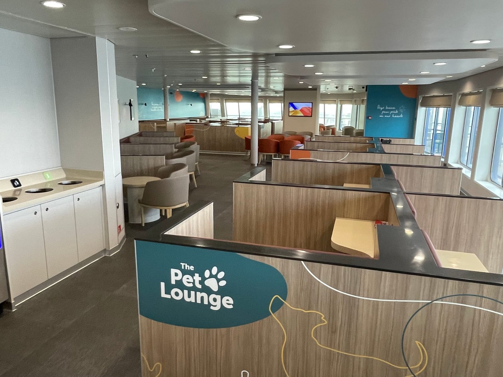 The Pet Lounge aan boord: zitjes met houten schotten ertussen, stoelen en ronde tafels, petrolblauwe wanden met de tekst Dogs leave paw prints on our hearts en ramen aan de zijkant