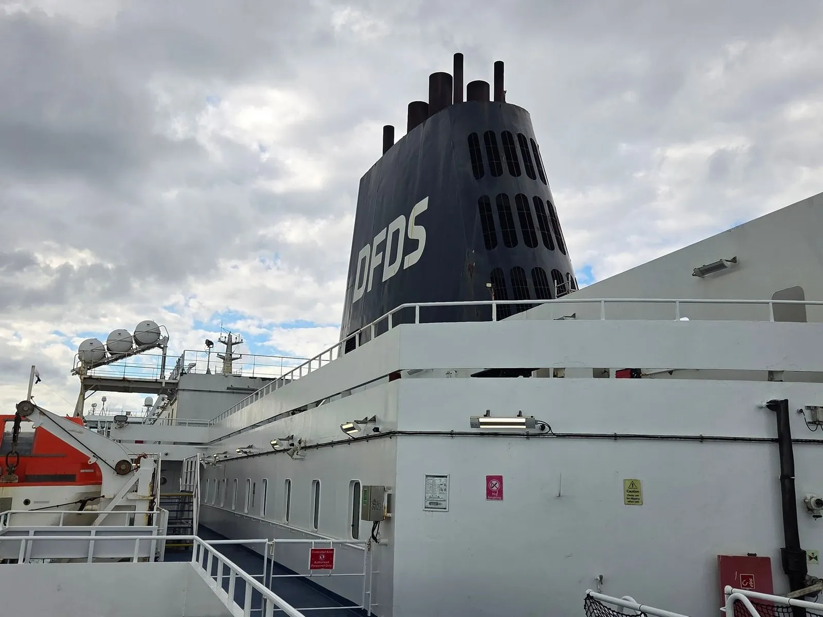 De zwarte DFDS-schoorsteen en het buitendek van de Delft Seaways onder een bewolkte lucht, met de reddingsboten ernaast