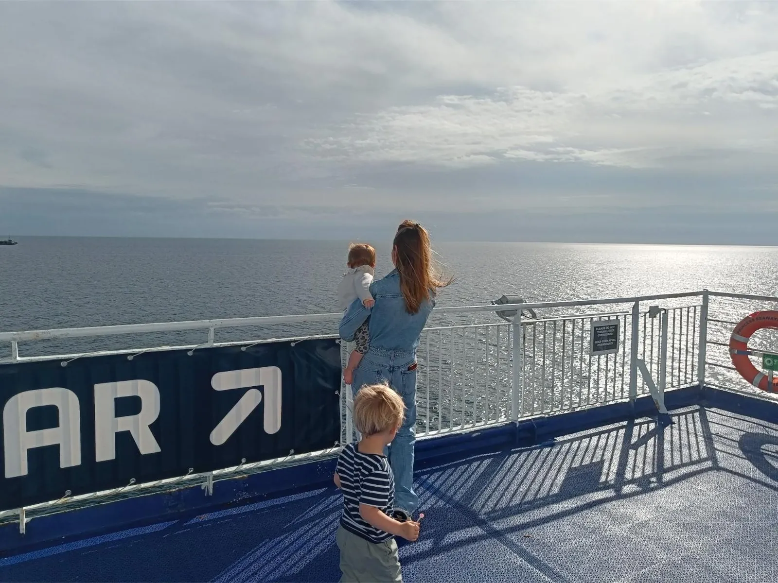Een volwassene en twee kinderen kijken vanaf het buitendek over zee, naast een bord dat naar de Sky Bar wijst