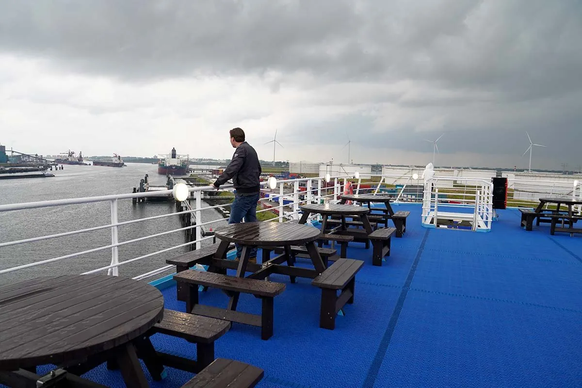 Het open bovendek met een blauwe vloer en houten picknicktafels; iemand staat aan de reling en kijkt over het water van het havengebied, met een tanker en windmolens in de verte