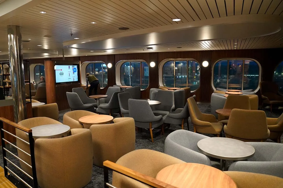 Zitgedeelte aan boord met lage fauteuils in grijs en okergeel rond kleine tafels, langs ovale ramen